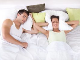 Ein Mann schläft schnarchend während seine Partnerin sich aus Frustration das Kissen gegen die Ohren drückt.