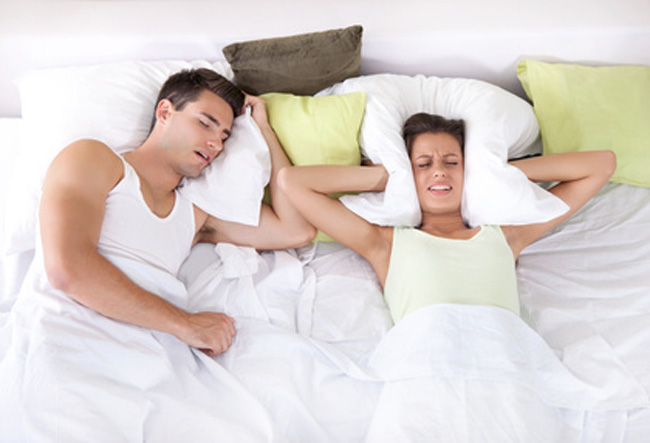 Ein Mann schläft schnarchend während seine Partnerin sich aus Frustration das Kissen gegen die Ohren drückt.