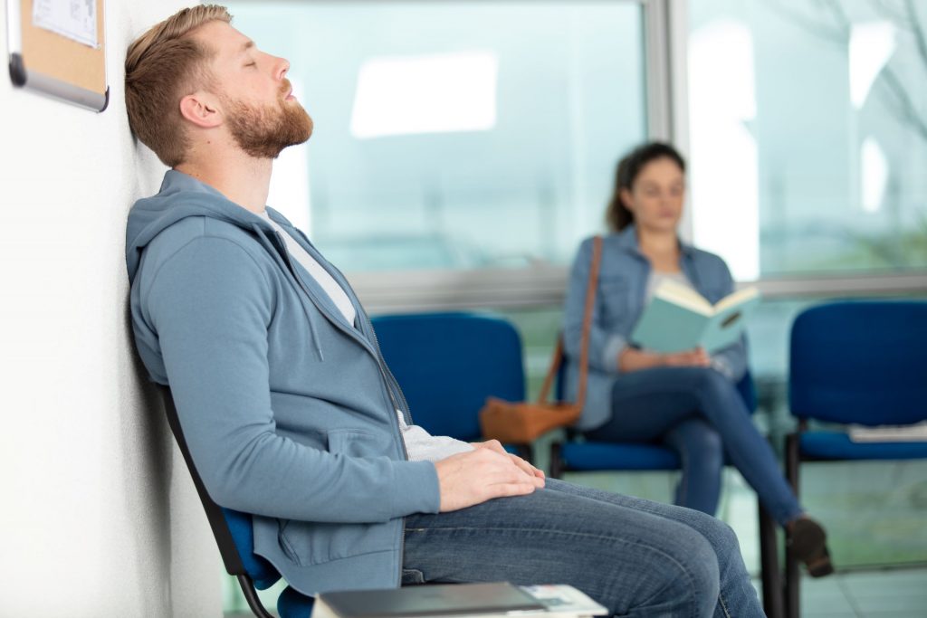 Erschöpfter Mann sitzt im Wartezimmer einer Arztpraxis und lehnt mit geschlossenen Augen an der Wand, während im Hintergrund eine Frau liest – Symbol für lange Wartezeiten auf einen Arzttermin.