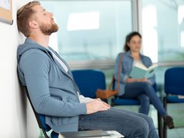 Erschöpfter Mann sitzt im Wartezimmer einer Arztpraxis und lehnt mit geschlossenen Augen an der Wand, während im Hintergrund eine Frau liest – Symbol für lange Wartezeiten auf einen Arzttermin.
