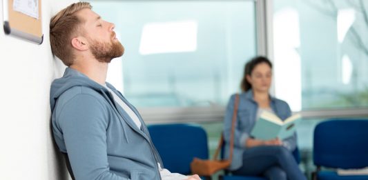 Ohne lange Wartezeiten einen Arzttermin erhalten: So ist es möglich Erschöpfter Mann sitzt im Wartezimmer einer Arztpraxis und lehnt mit geschlossenen Augen an der Wand, während im Hintergrund eine Frau liest – Symbol für lange Wartezeiten auf einen Arzttermin.