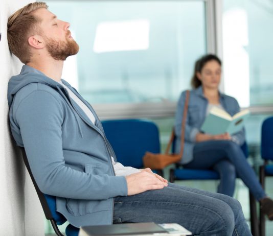 Erschöpfter Mann sitzt im Wartezimmer einer Arztpraxis und lehnt mit geschlossenen Augen an der Wand, während im Hintergrund eine Frau liest – Symbol für lange Wartezeiten auf einen Arzttermin.