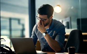 Ein Mann sitzt gestresst am Schreibtisch vor seinem Laptop und reibt sich die Augen – Symbolbild für beruflichen Stress, Überlastung und erhöhte Cortisolwerte durch dauerhafte Anspannung.