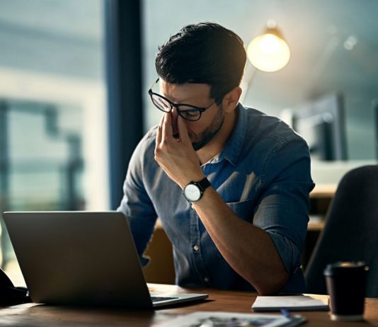 Ein Mann sitzt gestresst am Schreibtisch vor seinem Laptop und reibt sich die Augen – Symbolbild für beruflichen Stress, Überlastung und erhöhte Cortisolwerte durch dauerhafte Anspannung.