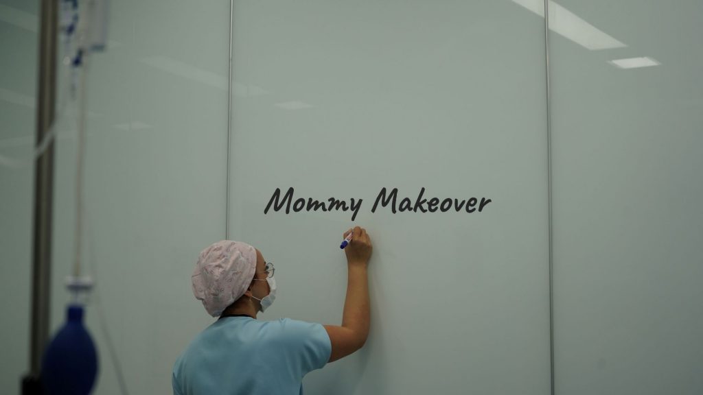Ärztin in OP-Kleidung und Mundschutz schreibt „Mommy Makeover“ auf eine weiße Wand in einer Praxis – Symbolbild für ästhetische Chirurgie nach Schwangerschaft.