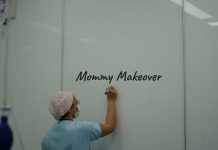 Ärztin in OP-Kleidung und Mundschutz schreibt „Mommy Makeover“ auf eine weiße Wand in einer Praxis – Symbolbild für ästhetische Chirurgie nach Schwangerschaft.