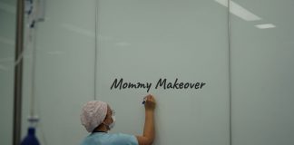 Dr. Frank Sander von Team Sander: „Das Mommy Makeover steht für einen bewussten Schritt hin zu mehr Selbstakzeptanz“ Ärztin in OP-Kleidung und Mundschutz schreibt „Mommy Makeover“ auf eine weiße Wand in einer Praxis – Symbolbild für ästhetische Chirurgie nach Schwangerschaft.