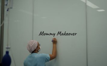 Ärztin in OP-Kleidung und Mundschutz schreibt „Mommy Makeover“ auf eine weiße Wand in einer Praxis – Symbolbild für ästhetische Chirurgie nach Schwangerschaft.