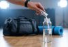 Welches Elektrolyt sollte ich kaufen? Hand gibt Elektrolytpulver in eine Wasserflasche vor dem Sport. Im Hintergrund liegen Sporttasche und Trainingsmatte. Symbolbild für Elektrolytgetränke, Mineralstoffausgleich und Regeneration nach körperlicher Belastung.