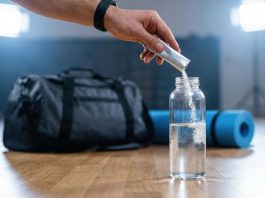 Hand gibt Elektrolytpulver in eine Wasserflasche vor dem Sport. Im Hintergrund liegen Sporttasche und Trainingsmatte. Symbolbild für Elektrolytgetränke, Mineralstoffausgleich und Regeneration nach körperlicher Belastung.