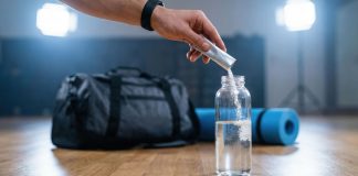 Hand gibt Elektrolytpulver in eine Wasserflasche vor dem Sport. Im Hintergrund liegen Sporttasche und Trainingsmatte. Symbolbild für Elektrolytgetränke, Mineralstoffausgleich und Regeneration nach körperlicher Belastung.