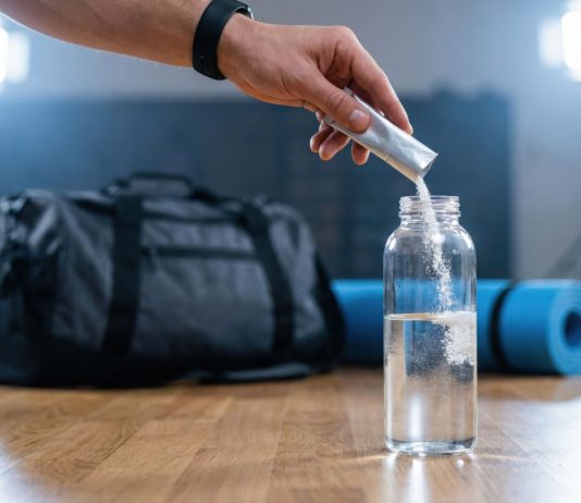 Hand gibt Elektrolytpulver in eine Wasserflasche vor dem Sport. Im Hintergrund liegen Sporttasche und Trainingsmatte. Symbolbild für Elektrolytgetränke, Mineralstoffausgleich und Regeneration nach körperlicher Belastung.