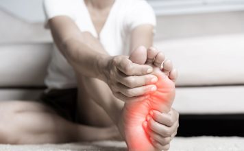 Person sitzt auf dem Boden und hält schmerzhaft die Fußsohle, der Bereich des Vorfußes ist rot markiert – Symbolbild für Fußschmerzen durch Fehlstellungen wie Spreiz- oder Plattfuß.