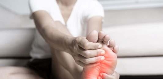 Person sitzt auf dem Boden und hält schmerzhaft die Fußsohle, der Bereich des Vorfußes ist rot markiert – Symbolbild für Fußschmerzen durch Fehlstellungen wie Spreiz- oder Plattfuß.