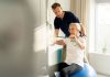 Hausbesuche und Praxisphysiotherapie: Was Ärzte ihren Patienten empfehlen sollten Physiotherapeut unterstützt ältere Patientin beim Training mit Widerstandsband auf einem Gymnastikball in einer hellen Praxis.