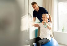 Hausbesuche und Praxisphysiotherapie: Was Ärzte ihren Patienten empfehlen sollten Physiotherapeut unterstützt ältere Patientin beim Training mit Widerstandsband auf einem Gymnastikball in einer hellen Praxis.