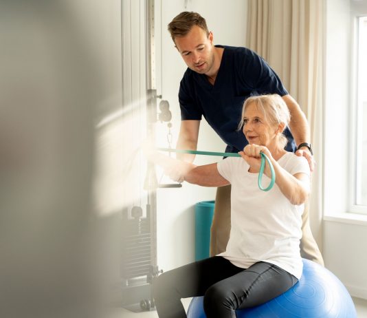 Physiotherapeut unterstützt ältere Patientin beim Training mit Widerstandsband auf einem Gymnastikball in einer hellen Praxis.