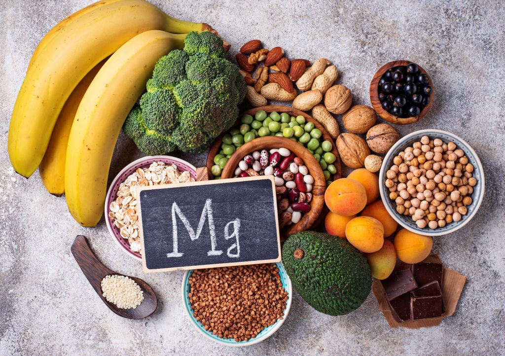 Magnesiumreiche Lebensmittel wie Bananen, Nüsse, Hülsenfrüchte, Brokkoli und dunkle Schokolade liegen neben einer Tafel mit Mg-Symbol.