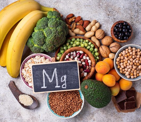 Magnesiumreiche Lebensmittel wie Bananen, Nüsse, Hülsenfrüchte, Brokkoli und dunkle Schokolade liegen neben einer Tafel mit Mg-Symbol.