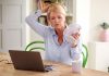 Ältere Frau kühlt sich während der Arbeit am Laptop mit einem Handventilator ab – typisches Bild für Hitzewallungen als Symptom der Wechseljahre bzw. Menopause.