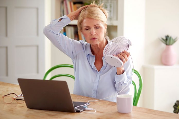 Ältere Frau kühlt sich während der Arbeit am Laptop mit einem Handventilator ab – typisches Bild für Hitzewallungen als Symptom der Wechseljahre bzw. Menopause.