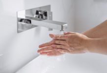 Hände werden unter fließendem Wasser an einem modernen, wandmontierten Wasserhahn gewaschen, Nahaufnahme im hellen Waschbeckenbereich, Fokus auf Hygiene und saubere Handreinigung.