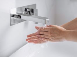 Hände werden unter fließendem Wasser an einem modernen, wandmontierten Wasserhahn gewaschen, Nahaufnahme im hellen Waschbeckenbereich, Fokus auf Hygiene und saubere Handreinigung.