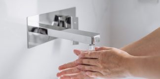 Hände werden unter fließendem Wasser an einem modernen, wandmontierten Wasserhahn gewaschen, Nahaufnahme im hellen Waschbeckenbereich, Fokus auf Hygiene und saubere Handreinigung.