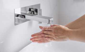 Hände werden unter fließendem Wasser an einem modernen, wandmontierten Wasserhahn gewaschen, Nahaufnahme im hellen Waschbeckenbereich, Fokus auf Hygiene und saubere Handreinigung.