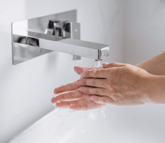 Hände werden unter fließendem Wasser an einem modernen, wandmontierten Wasserhahn gewaschen, Nahaufnahme im hellen Waschbeckenbereich, Fokus auf Hygiene und saubere Handreinigung.