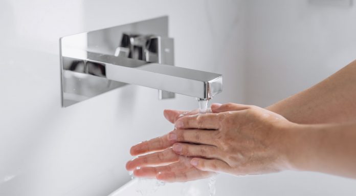 Hände werden unter fließendem Wasser an einem modernen, wandmontierten Wasserhahn gewaschen, Nahaufnahme im hellen Waschbeckenbereich, Fokus auf Hygiene und saubere Handreinigung.