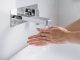 Hände werden unter fließendem Wasser an einem modernen, wandmontierten Wasserhahn gewaschen, Nahaufnahme im hellen Waschbeckenbereich, Fokus auf Hygiene und saubere Handreinigung.