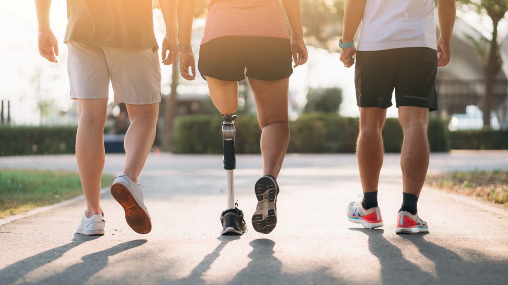 Drei Menschen joggen bei warmem Sonnenlicht auf einem Parkweg; die Person in der Mitte läuft mit einer Unterschenkelprothese.