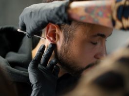 Mann bekommt Piercing am Ohrläppchen, Piercer trägt mit Handschuhen antiseptische Lösung auf – hygienische Vorbereitung vor dem Stechen eines Ohrpiercings in einem Piercingstudio.