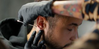 Mann bekommt Piercing am Ohrläppchen, Piercer trägt mit Handschuhen antiseptische Lösung auf – hygienische Vorbereitung vor dem Stechen eines Ohrpiercings in einem Piercingstudio.