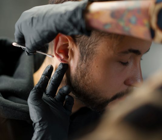 Mann bekommt Piercing am Ohrläppchen, Piercer trägt mit Handschuhen antiseptische Lösung auf – hygienische Vorbereitung vor dem Stechen eines Ohrpiercings in einem Piercingstudio.
