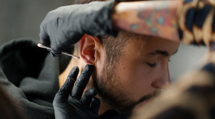 Mann bekommt Piercing am Ohrläppchen, Piercer trägt mit Handschuhen antiseptische Lösung auf – hygienische Vorbereitung vor dem Stechen eines Ohrpiercings in einem Piercingstudio.