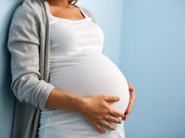 Eine schwangere Frau lehnt an einer Wand und legt ihre Hände auf ihren Bauch.