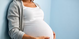 Eine schwangere Frau lehnt an einer Wand und legt ihre Hände auf ihren Bauch.