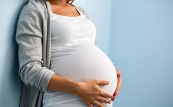 Eine schwangere Frau lehnt an einer Wand und legt ihre Hände auf ihren Bauch.