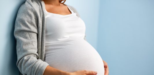 Eine schwangere Frau lehnt an einer Wand und legt ihre Hände auf ihren Bauch.