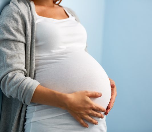 Eine schwangere Frau lehnt an einer Wand und legt ihre Hände auf ihren Bauch.