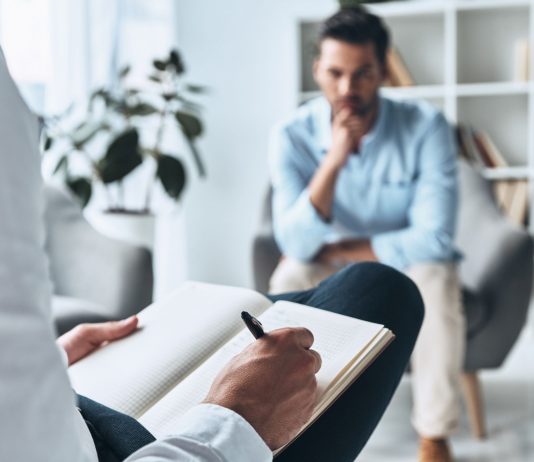 Psychotherapeutische Sitzung: Ein Mann spricht mit einem Therapeuten, der Notizen macht. Thema sind Trauma, Angststörungen und moderne Traumatherapie in ruhiger Praxisumgebung.