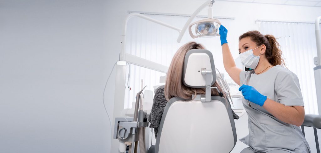 Zahnärztin mit Maske und Handschuhen bereitet in einer hellen Praxis die zahnärztliche Untersuchung einer Patientin auf dem Behandlungsstuhl vor.