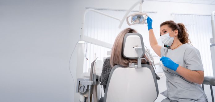 Zahnärztin mit Maske und Handschuhen bereitet in einer hellen Praxis die zahnärztliche Untersuchung einer Patientin auf dem Behandlungsstuhl vor.